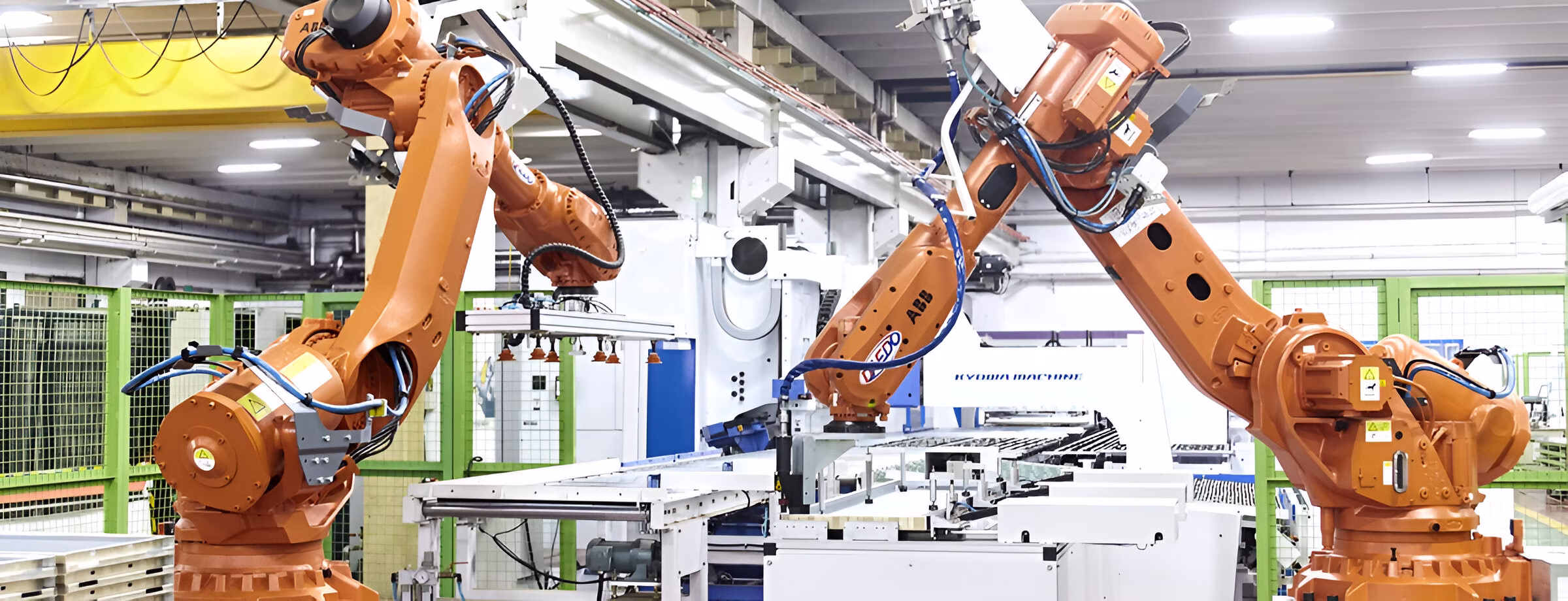 Зображення демонструє виробничий цех з двома великими помаранчевими промисловими роботами-маніпуляторами типу ABB, розташованими обабіч автоматизованої конвеєрної лінії. Роботи встановлені у світлому приміщенні із зеленими захисними огорожами на задньому плані. До роботів підключені різноманітні кабелі та пневматичні лінії. Ця система представляє сучасну автоматизовану технологію, яка використовується в автомобільній, електронній та інших галузях промисловості.
