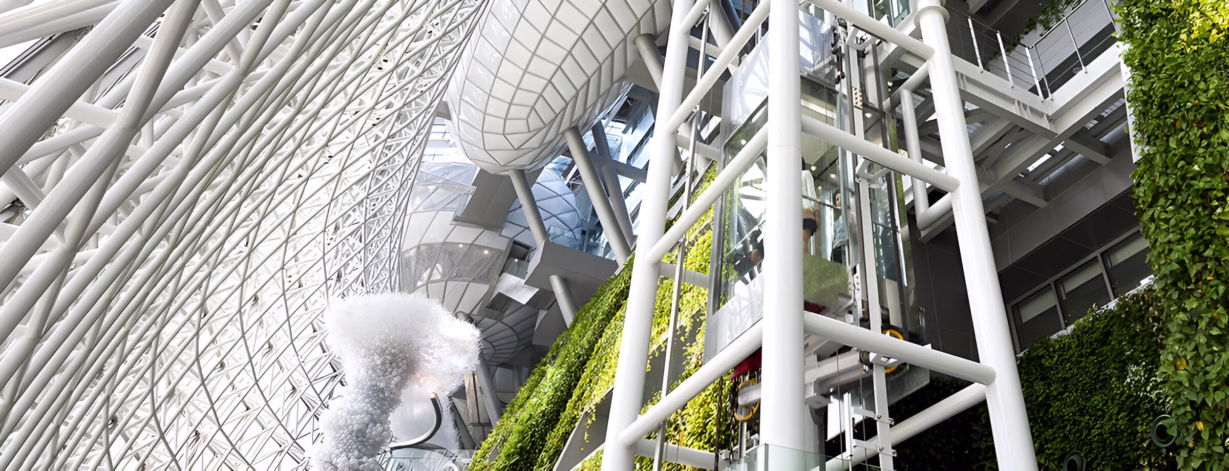 Зображення демонструє сучасний архітектурний простір із панорамним скляним ліфтом, розташованим у металевій конструкції. Інтер'єр поєднує хай-тек дизайн із природними елементами, зокрема вертикальними зеленими стінами. Стеля виконана у вигляді складної геометричної решітки, що пропускає багато природного світла. Це може бути частина бізнес-центру, готелю чи громадської будівлі з футуристичним дизайном.