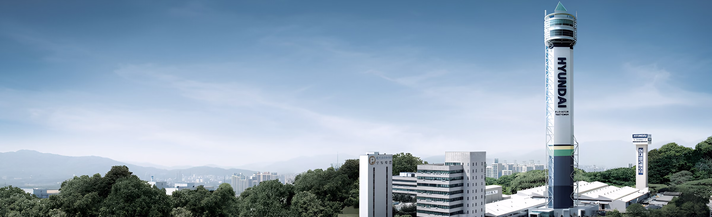Комплекс штаб-квартири Hyundai з характерною високою вежею, на якій вертикально написано 'HYUNDAI'. На вершині вежі є скляна оглядова кімната із зеленою пірамідальною верхівкою. Комплекс включає кілька офісних будівель, оточених зеленню. На задньому плані видно міський пейзаж з горами на горизонті під блакитним небом.