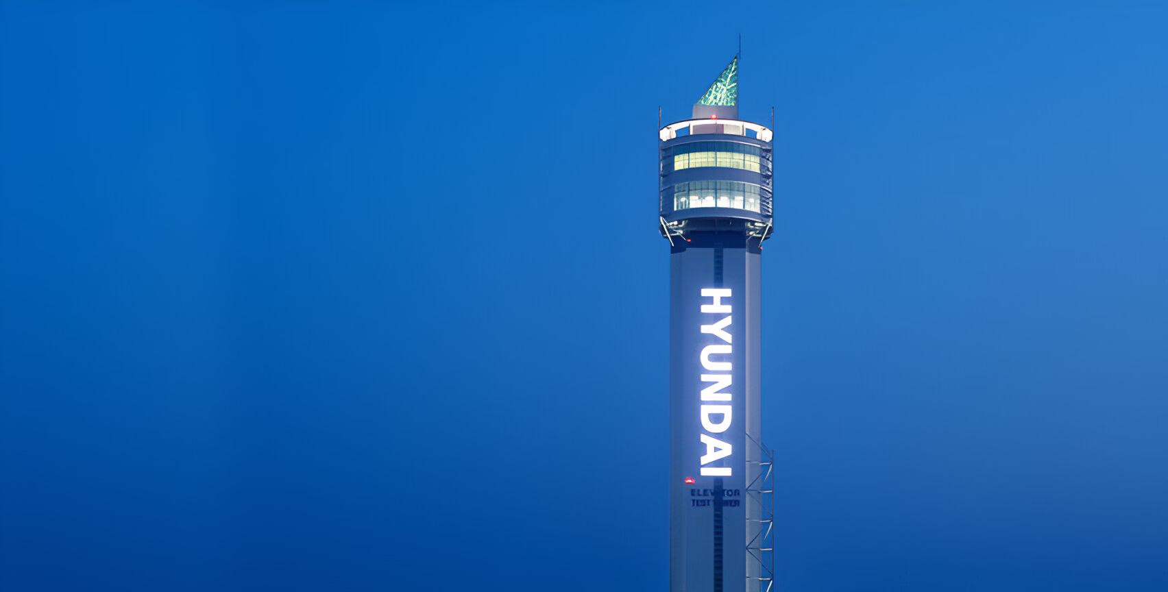 Висока вежа Hyundai Elevator з підсвіченою назвою компанії вертикально вздовж її стовбура на тлі темно-синього вечірнього неба.
