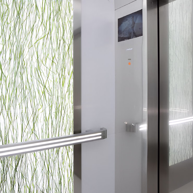 Фрагмент інтер'єру ліфта з сучасним дизайном: видно частину дверей ліфта зі срібною ручкою та панель керування з невеликим дисплеєм. Стіни декоровані напівпрозорими панелями з елегантним зеленим візерунком, що нагадує стебла рослин. Весь дизайн створює легке, природне відчуття в сучасному технологічному просторі.