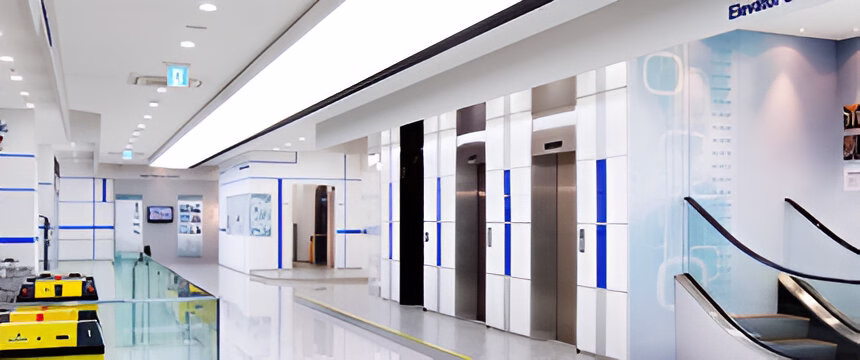 Сучасне просторе фойє бізнес-центру або торгового комплексу з ліфтовою зоною. На фото видно кілька ліфтів із металевими та білими дверима з синіми акцентними смугами. Інтер'єр виконаний переважно в білому кольорі з голубими елементами. Ліворуч видно скляне огородження та частину ескалатора праворуч. Стеля оснащена вбудованими світильниками. У приміщенні чисто, світло та виглядає дуже сучасно, що створює атмосферу високотехнологічного простору.