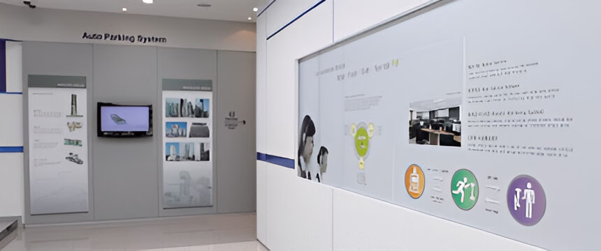 Виставковий зал із інформаційними стендами про системи автоматизації. Ліворуч видно експозицію з назвою 'Auto Pushing System' з екраном та зображеннями обладнання. Праворуч — біла інформаційна панель з різнокольоровими круглими іконками (жовта, оранжева, зелена, фіолетова), схемами та текстом. Приміщення оформлене в світлих кольорах з синіми акцентами. Стіни білі та сірі, стеля обладнана точковими світильниками. Загальний дизайн має сучасний корпоративний стиль, характерний для технологічних виставок.