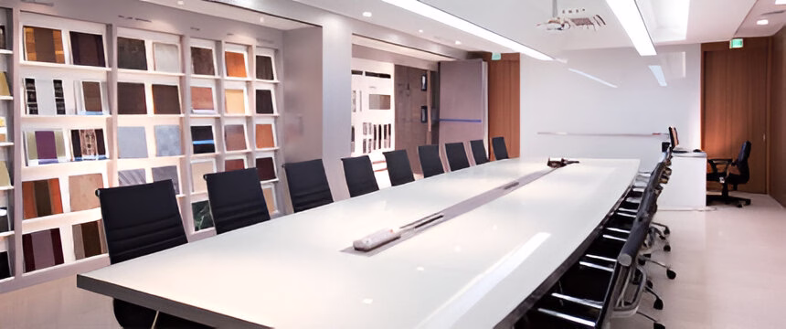 Сучасна конференц-зала зі світлим довгим столом, чорними кріслами, білими стінами та полицями з різноманітними зразками матеріалів на задньому плані.