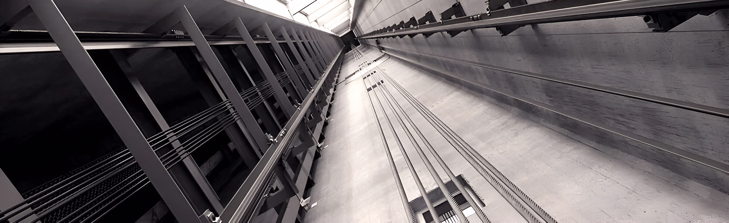 Чорно-біле зображення шахти ліфта, погляд знизу вгору, що демонструє металеві конструкції, напрямні рейки та троси, що тягнуться вгору до світлого отвору вгорі шахти.