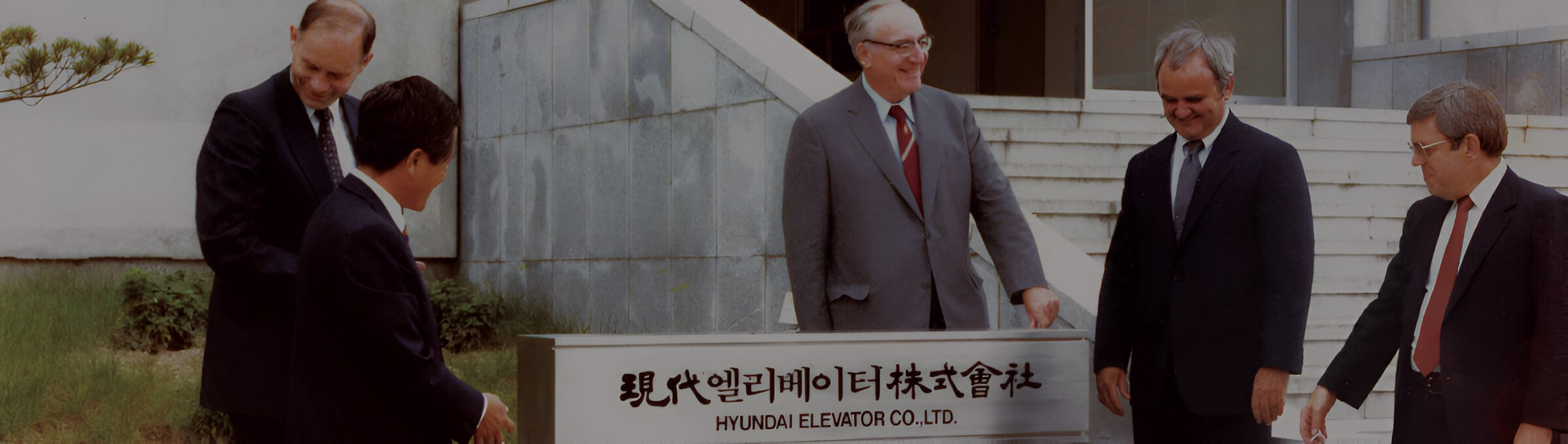 П'ятеро чоловіків у ділових костюмах стоять біля вивіски 'Hyundai Elevator Co., Ltd.' перед будівлею.