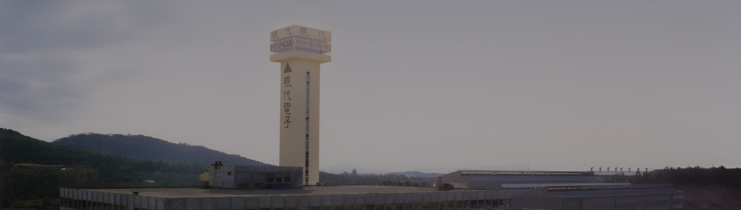 Висока біла вежа з написом китайською мовою на території промислового комплексу на фоні гір та хмарного неба.