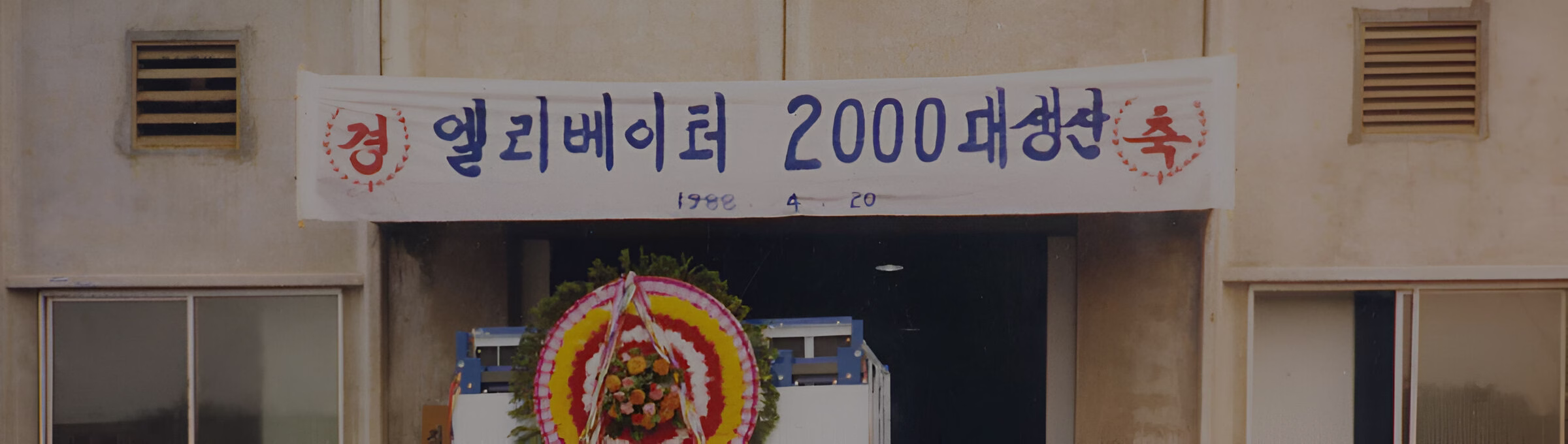 Будівля з білим банером над входом, на якому корейською мовою написано 'Святкування виробництва 2000 ліфтів Kyung', а також дата '1988 4 20', та великий квітковий вінок біля входу.