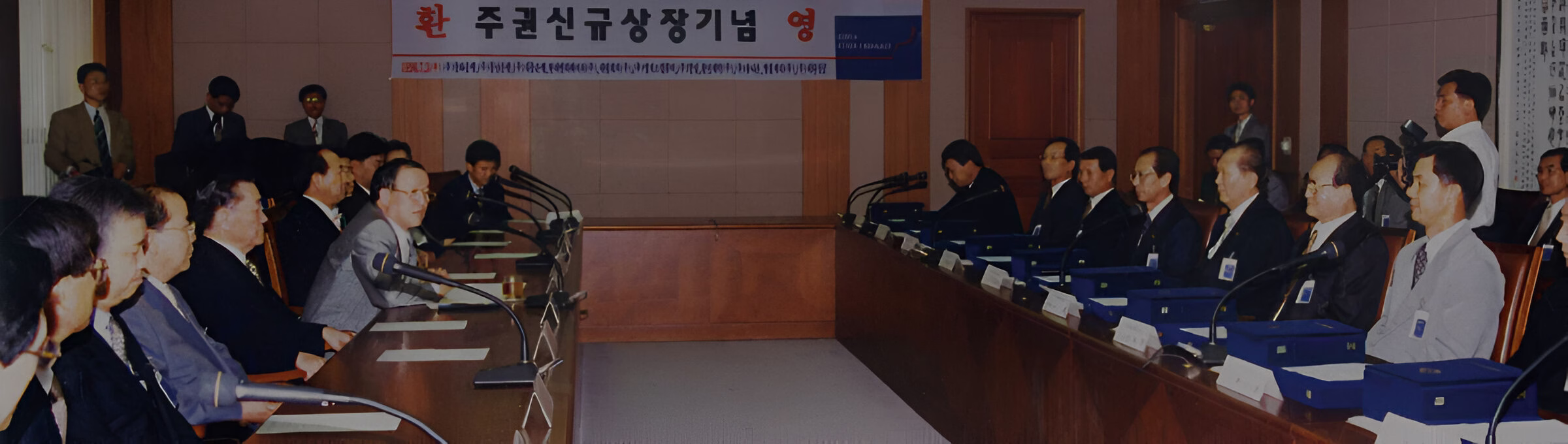 Група людей у формальному одязі зібралася в залі за довгими столами перед банером з корейським написом.