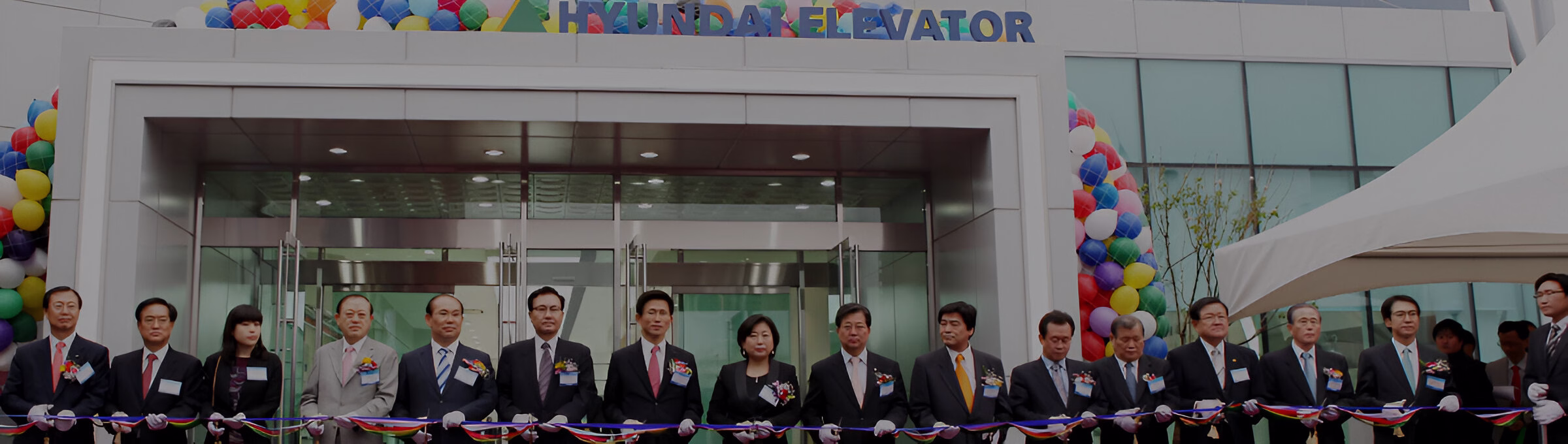 Урочисте відкриття будівлі Hyundai Elevator: офіційні особи та гості в ділових костюмах стоять в ряд за синьою стрічкою, тримаючи ножиці для її перерізання перед головним входом, прикрашеним арками з повітряних кульок.