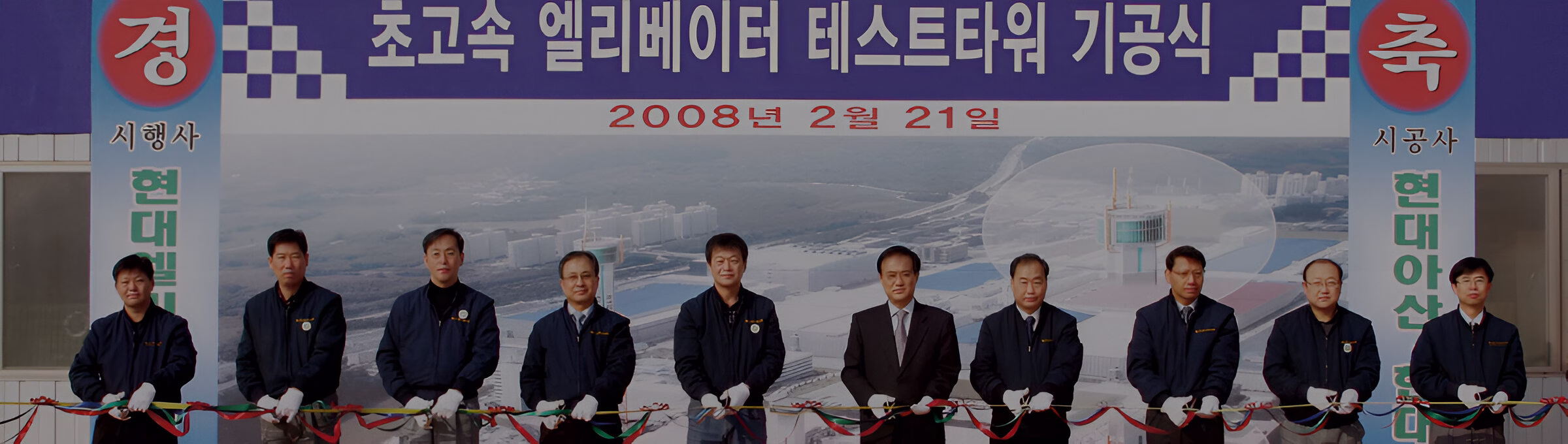 Дев'ять чоловіків у білих рукавичках стоять за церемоніальною стрічкою перед великим банером з написом корейською '초고속 엘리베이터 테스트 타워 기공식' та датою '2008년 2월 21일', що позначає церемонію початку будівництва тестової вежі.