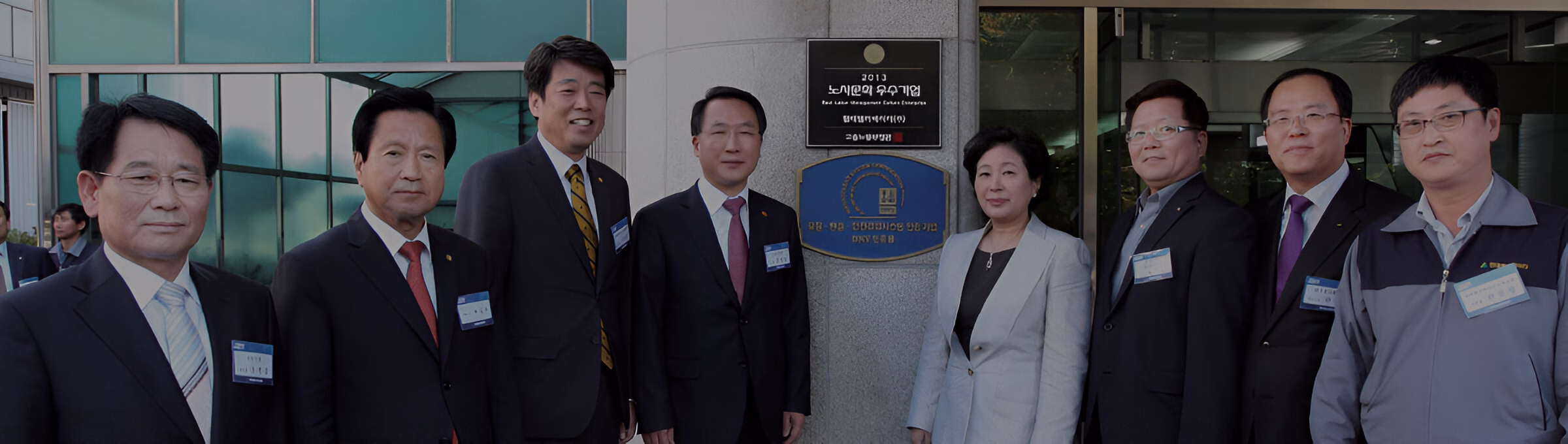 Офіційне групове фото: семеро чоловіків та одна жінка у діловому одязі позують на вулиці перед входом у будівлю, поруч із яким на стіні висить табличка з написом (частково видно '2013' та корейські символи).
