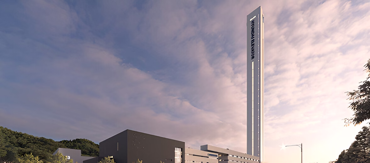 Сучасна архітектурна композиція, що включає високу вертикальну вежу з написом на ній, та кілька низьких будівель поруч. На задньому плані видно пагорби з зеленою рослинністю. Небо частково вкрите хмарами, створюючи ніжний світловий ефект. Загальний вигляд нагадує кампус чи діловий центр в оточенні природи.