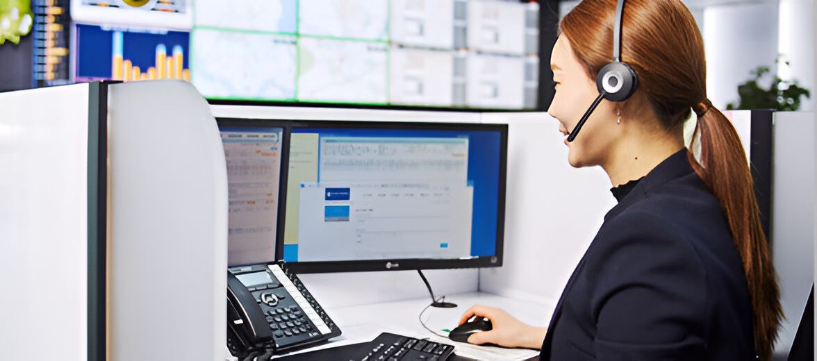 Жінка з рудим волоссям, зібраним у хвіст, працює за комп'ютером у навушниках з мікрофоном. Вона сидить у профіль в офісному просторі перед монітором з відкритими вікнами програм і даними. На столі видно телефон та комп'ютерну мишу. На задньому плані розташовані екрани з графіками та діаграмами, що показують різну статистику. Робоче місце виглядає як центр обробки викликів або моніторингова станція.