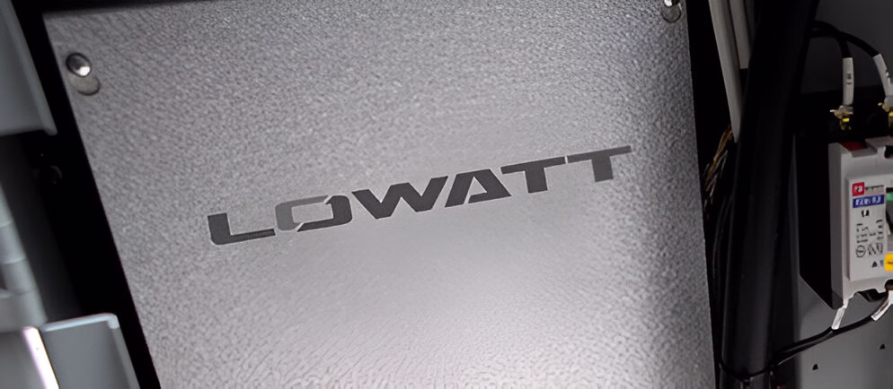 Крупний план металевої панелі сірого кольору з вирізаним великим написом 'LOWATT', встановленої серед іншого обладнання з кабелями та електричними компонентами.