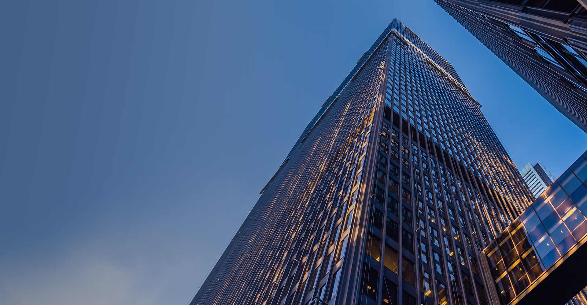 Високий сучасний хмарочос із унікальним дизайном фасаду, що поєднує скляні панелі з відблисками синього та золотистого кольорів. Будівля височіє на тлі яскравого блакитного неба з білими хмарами.