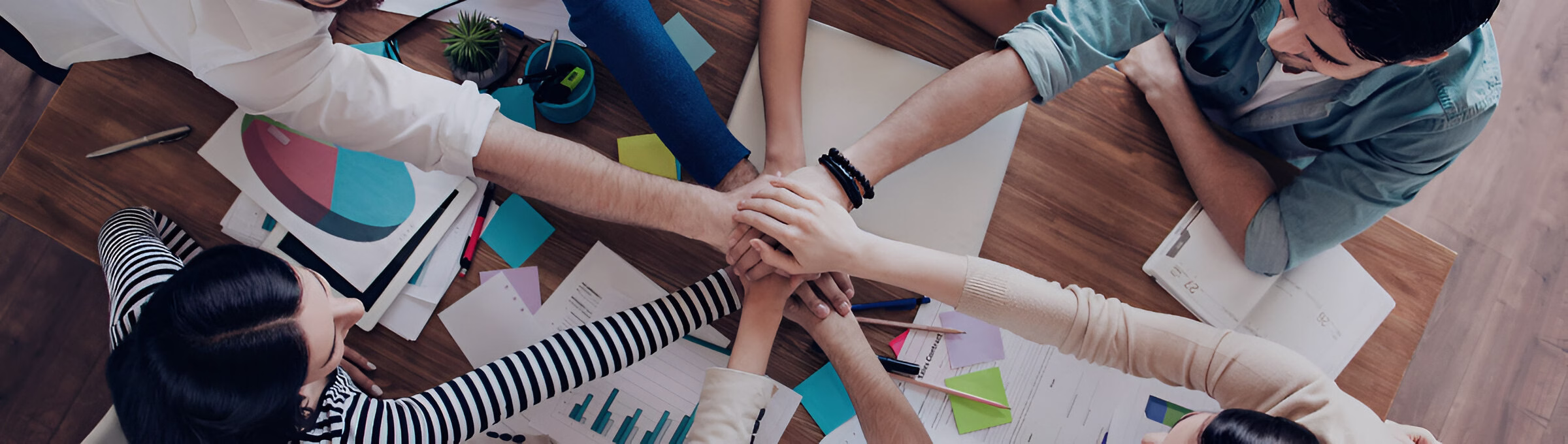 Вигляд зверху на робочий стіл, де четверо людей кладуть руки одна на одну в центрі столу, демонструючи командну єдність та співпрацю. На столі розкладені документи з графіками, кольорові стікери та канцелярське приладдя, що свідчить про спільну роботу над проєктом або бізнес-планом.