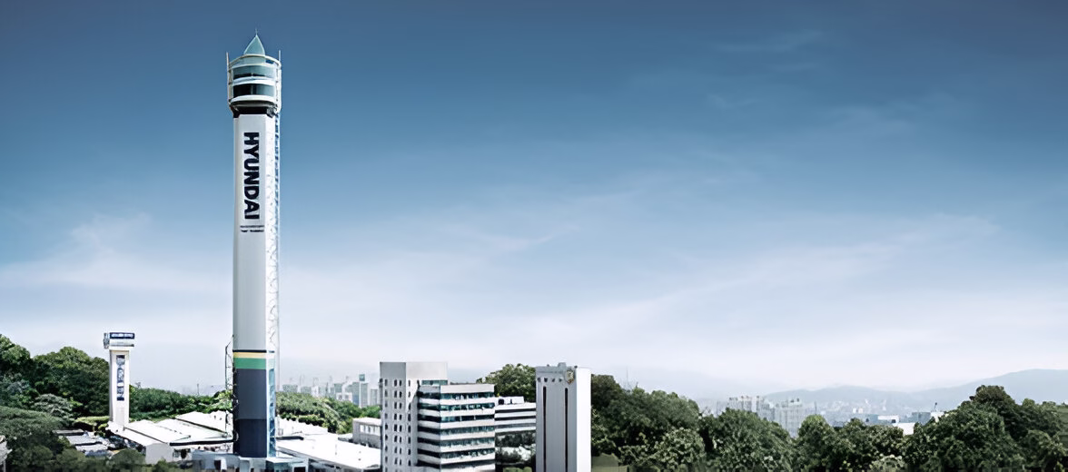 Висока біла вежа з написом 'HYUNDAI' вертикально вздовж її сторони, розташована серед міської забудови та зелених насаджень під блакитним небом. На вершині вежі знаходиться оглядовий майданчик зі скляними стінами та конічним дахом. Ще одна менша вежа з написом 'HYUNDAI' видніється ліворуч на задньому плані.