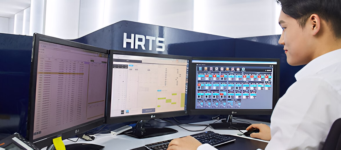 Чоловік азіатської зовнішності сидить за робочим столом перед трьома комп'ютерними моніторами, на яких відображаються різні інтерфейси програм. Він одягнений у світлу сорочку та працює з клавіатурою та мишкою. На задньому плані видно темну панель з написом 'HRTS'.