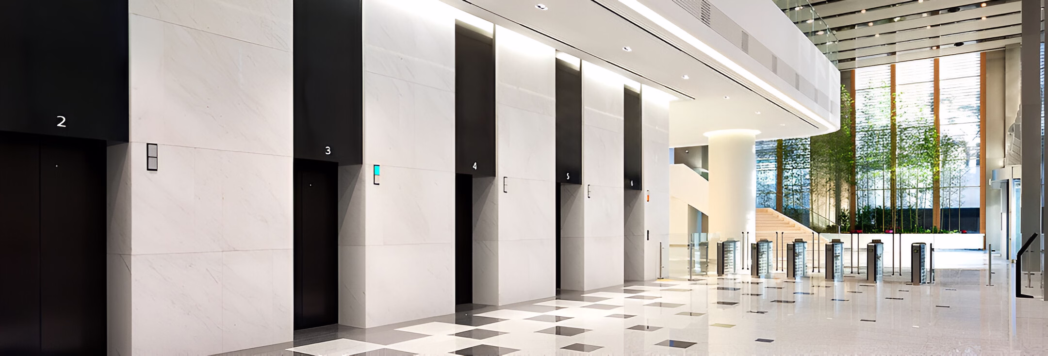 Сучасний та просторий інтер'єр будівлі з рядом чорних ліфтових дверей, пронумерованих 2, 3, 4, 5 та 6, розташованих вздовж стіни, оздобленої світлим каменем з чорними вертикальними вставками. Підлога вкрита світлою плиткою з геометричним візерунком. У дальньому кінці видно турнікети та скляні стіни, за якими проглядають зелені насадження та сходи. Зверху приміщення освітлюється штучним світлом та частково природним світлом, що проникає через скляний фасад.