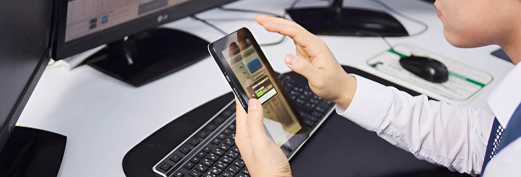 Людина в білій сорочці та краватці працює з планшетом, тримаючи його обома руками. На екрані планшета відображається інтерфейс програми з різними елементами керування. На задньому плані видно клавіатуру, комп'ютерну мишку та частину монітора на білому робочому столі.