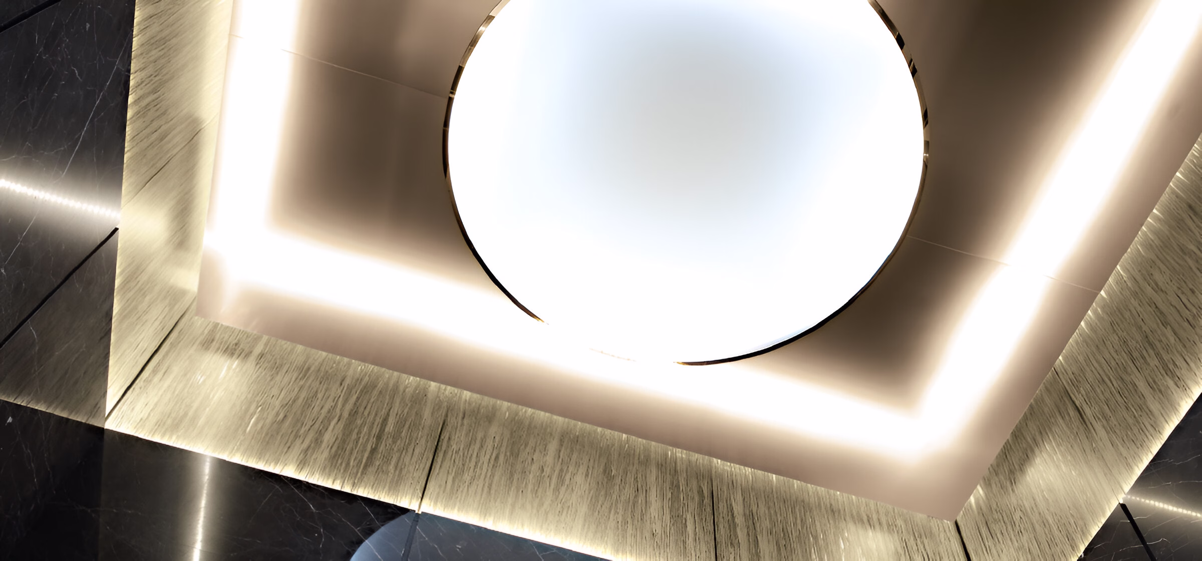 Стильний елемент інтер'єрного освітлення з круглою світловою панеллю на стелі, оточеною квадратною рамкою з підсвіткою. Стеля оздоблена текстурованими панелями світлого кольору, що контрастують із темними елементами інтер'єру. Світлодіодне підсвічування створює м'яке, розсіяне світло, підкреслюючи сучасний мінімалістичний дизайн приміщення.