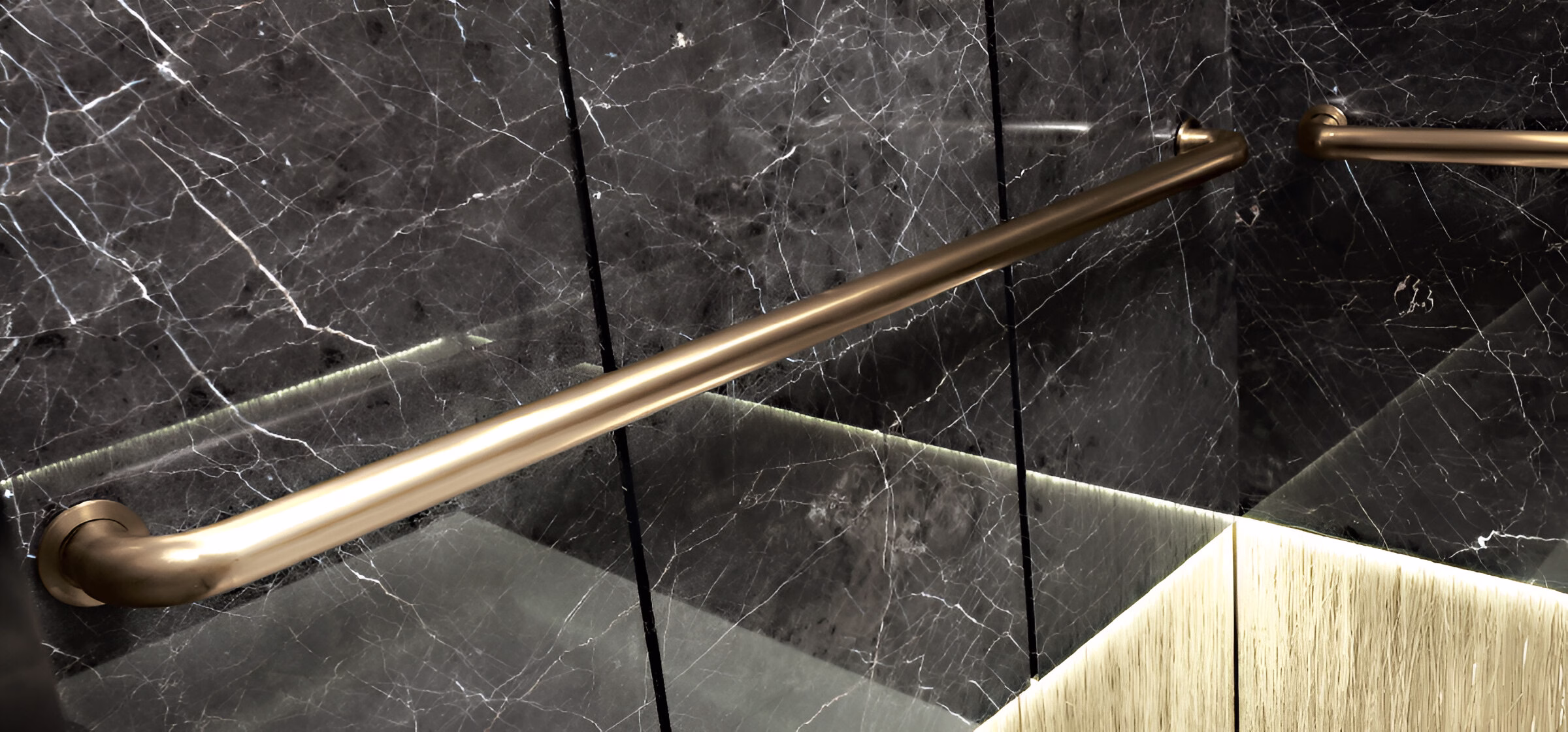 Елегантний латунний поручень на стіні з чорного мармуру. Поручень має матову золотисту поверхню і розташований горизонтально вздовж стіни. Мармур має характерні білі прожилки на темному тлі. У нижній частині зображення видно частину меблів зі світлою текстурою дерева та м'яке підсвічування. Дизайн інтер'єру виконаний у розкішному сучасному стилі з використанням преміальних матеріалів.