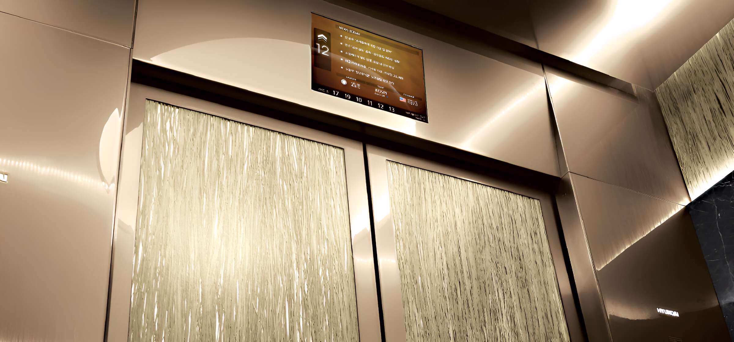 Сучасний інтер'єр ліфта з дизайнерським оздобленням. Стіни ліфта облицьовані панелями зі світлою вертикальною текстурою, обрамлені металевими елементами. Вгорі розташований цифровий інформаційний дисплей, що показує поточний поверх '12' та іншу інформацію. По периметру стелі ліфта встановлене м'яке приховане підсвічування, яке створює елегантну атмосферу. Дизайн поєднує функціональність із преміальною естетикою.