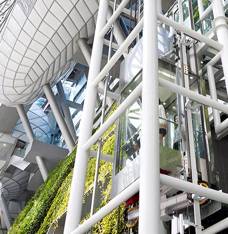 Вид знизу на сучасну будівлю з відкритими ліфтовими шахтами, скляними ліфтами, металевими конструкціями та вертикальним озелененням.