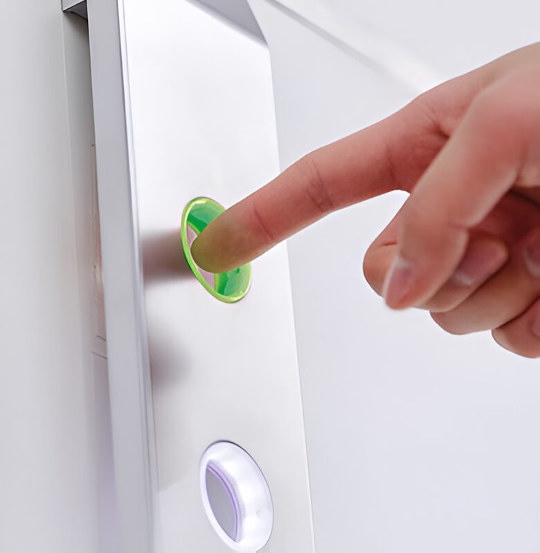 Палець натискає на зелену круглу кнопку з рожевим контуром на сріблястій панелі поруч з іншою білою підсвіченою кнопкою.