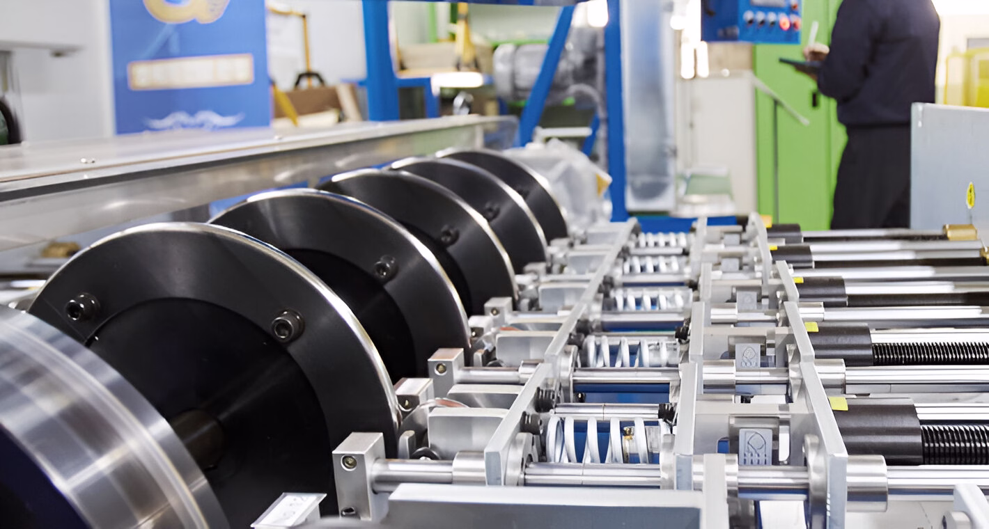 Промислова машина з великими металевими дисками та складною системою механізмів у виробничому цеху, на задньому плані видно силует людини біля панелі керування.