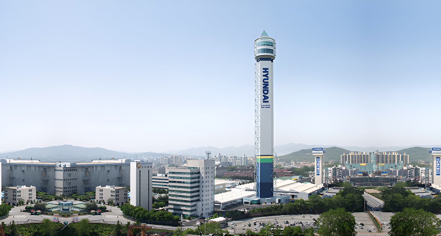 Панорамний вид на випробувальну вежу Hyundai Elevator, що височіє над промисловим комплексом серед зелених насаджень та гір на горизонті під ясним небом.