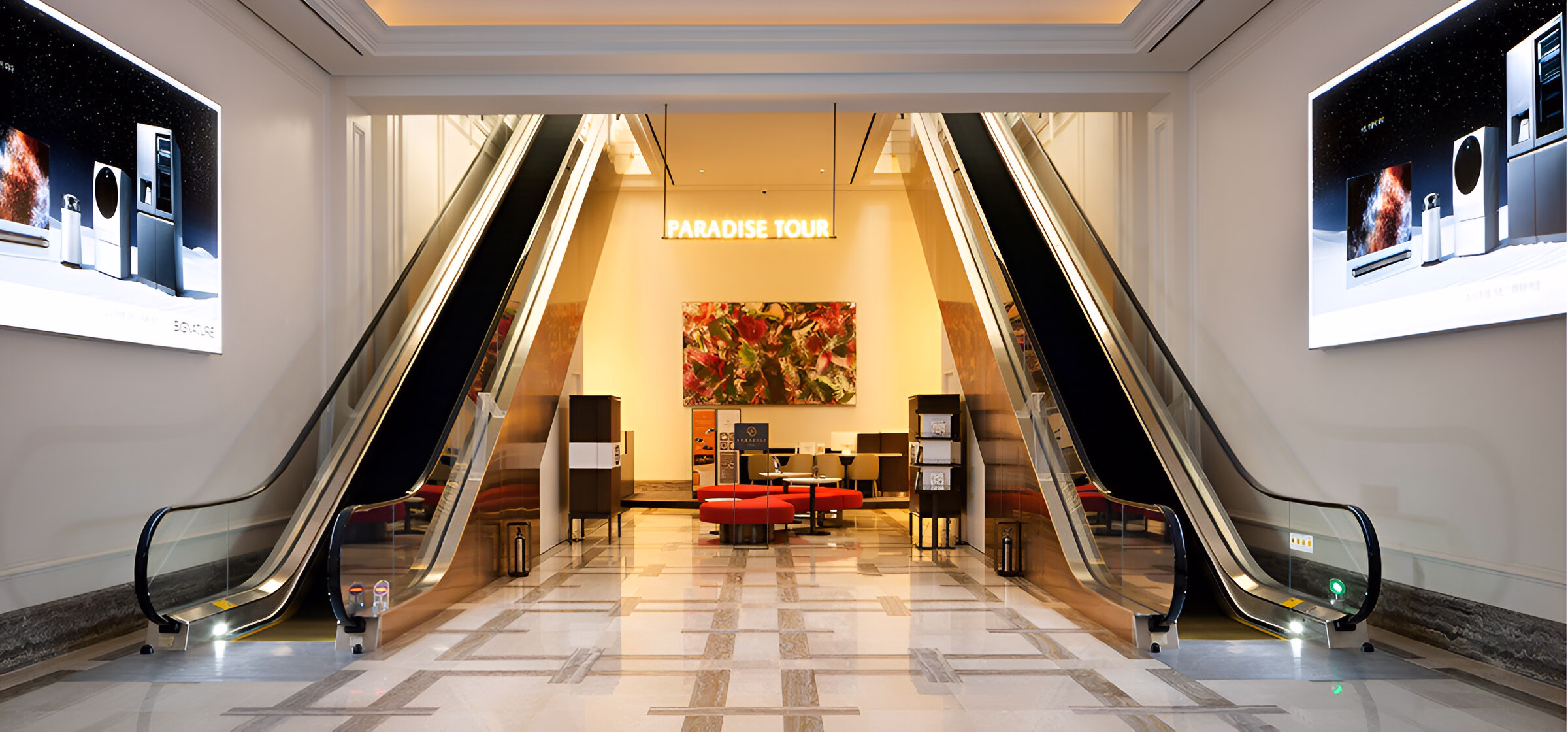 Інтер'єр сучасної будівлі з двома ескалаторами, що піднімаються вгору, між якими розташований прохід до зони відпочинку з червоними кріслами та столиками, а також картиною на стіні та написом 'PARADISE TOUR' вдалині, по боках ескалаторів розміщені рекламні екрани.