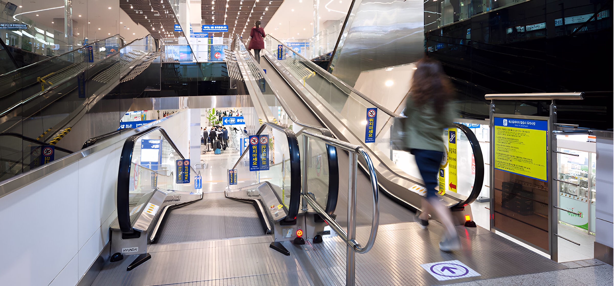 Вигляд зверху на пасажирів, які користуються ескалаторами в сучасному торговому центрі або транспортному терміналі з великою кількістю ескалаторів, покажчиками та скляними балюстрадами.