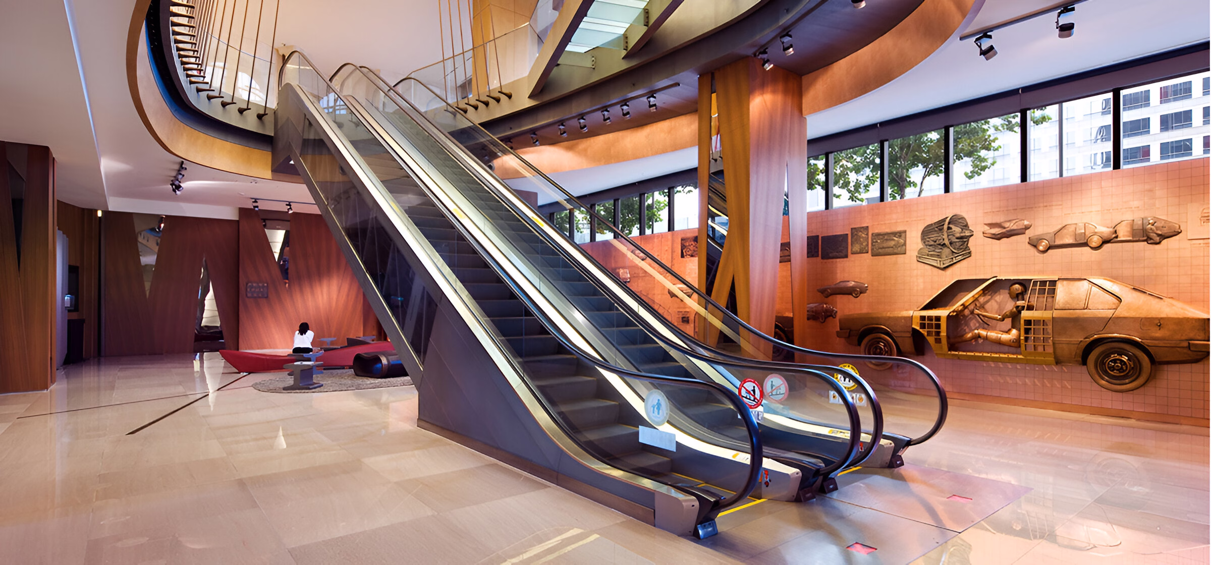 Інтер'єр сучасної будівлі з двома ескалаторами, що піднімаються вгору, розташованими поруч із зоною відпочинку з червоними диванами та журнальним столиком, а також стіною, прикрашеною дерев'яними барельєфами автомобілів та інших об'єктів.