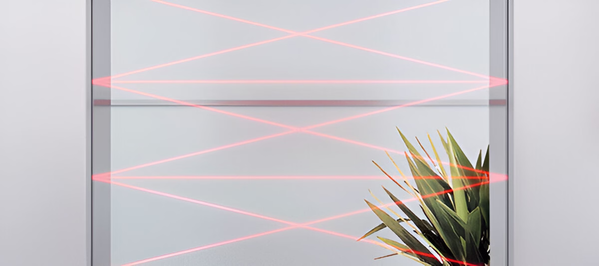 Червоні лазерні промені перетинають отвір ліфта, у нижньому правому куті видно зелену рослину, на тлі світлих стін.