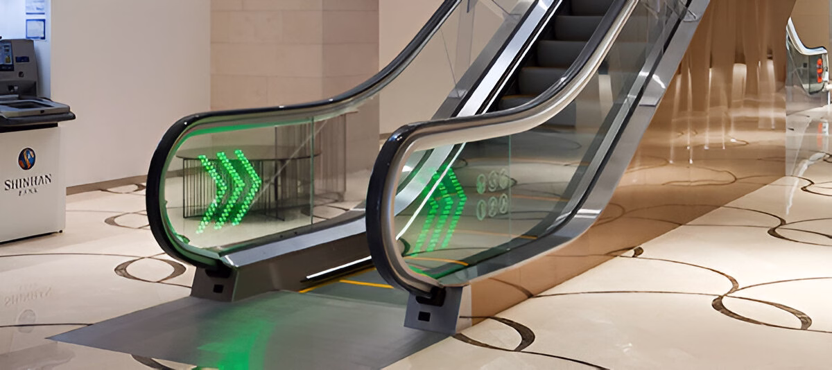 Початок ескалатора з прозорими балюстрадами, підсвіченими зеленими стрілками, що вказують напрямок руху вгору, на підлозі з візерунком, поруч із банкоматом або інформаційним кіоском.