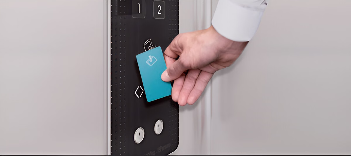 Чоловіча рука в білій сорочці вставляє блакитну картку з білим логотипом у чорний зчитувач карток, розташований вертикально поруч із кнопками поверхів '1' та '2' у ліфті зі світлими стінами.