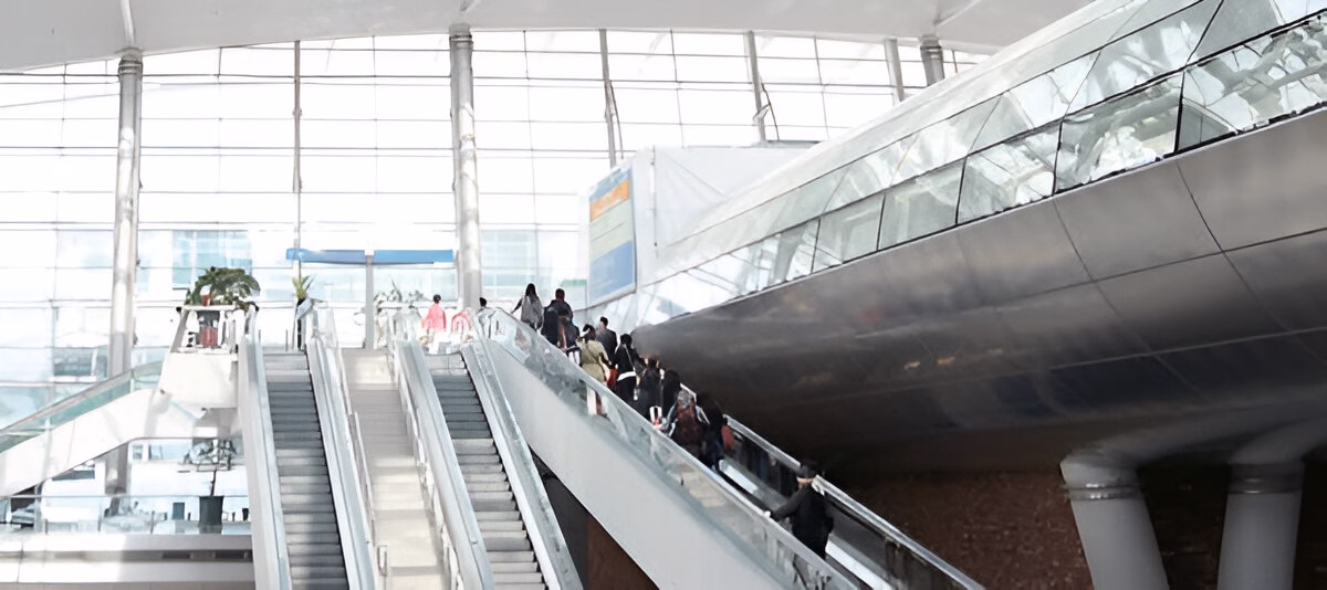 Пасажири з багажем піднімаються ескалаторами у світлому сучасному аеропорту з великими вікнами та металевими елементами конструкції.