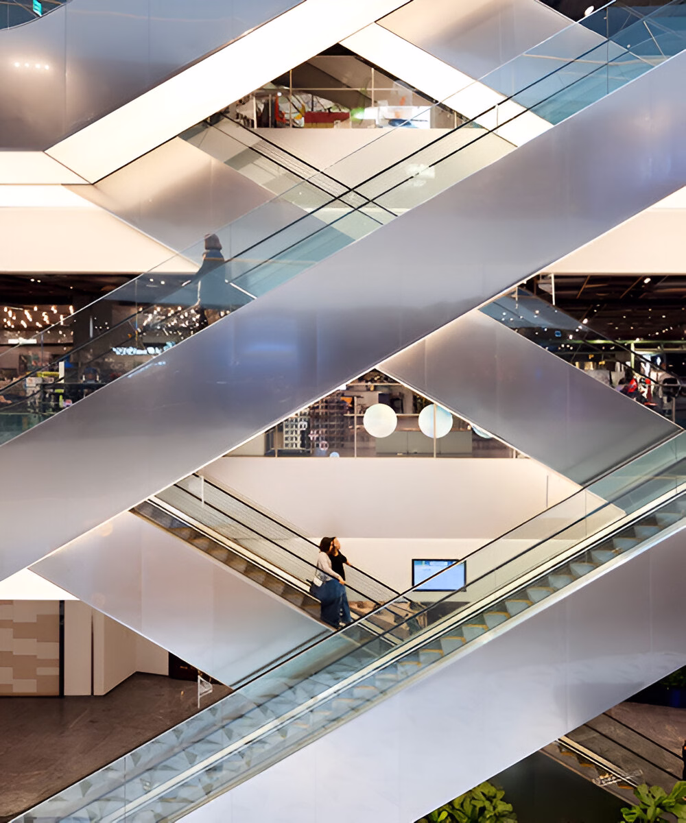 Багаторівневий інтер'єр торгового центру з перехресними ескалаторами, скляними балюстрадами та світлою обробкою, де люди переміщуються між поверхами, а внизу видно зелені рослини.